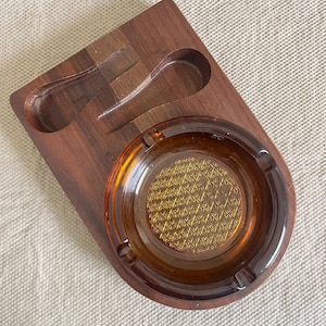 Könnte beinhalten: Ein Vintage-Aschenbecher und Pfeifenablage-Kombination. Der Aschenbecher besteht aus bernsteinfarbenem Glas mit strukturiertem Zentrum, eingebettet in eine dunkle Holzbasis. Die Basis hat zwei gebogene Rillen zum Ablegen von Pfeifen. Das Gesamtdesign ist rechteckig.