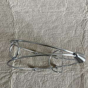 May include: A silver wine bottle holder with a spring handle and a circular design. The metal holder is designed to cradle a wine bottle. The holder is on a neutral-coloured fabric.