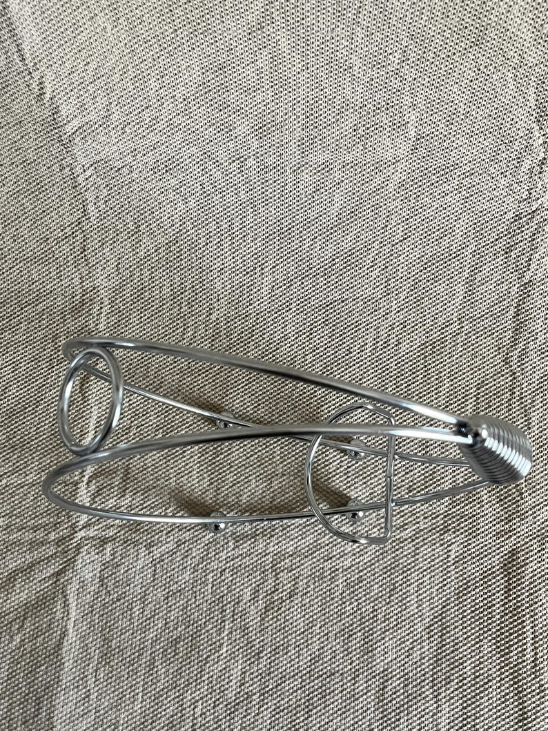 May include: A silver wine bottle holder with a spring handle and a circular design. The metal holder is designed to cradle a wine bottle. The holder is on a neutral-coloured fabric.