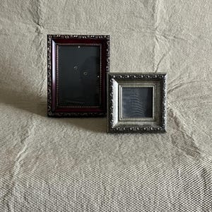 Puede incluir: Dos marcos de fotos decorativos. Uno es rectangular con un acabado burdeos oscuro y detalles ornamentados. El otro es cuadrado con un acabado plateado y detalles ornamentados similares. Ambos marcos tienen un interior oscuro.
