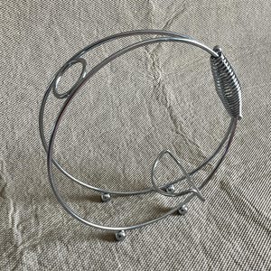 May include: A silver metal wine bottle holder with a circular design. It has a spring-like handle and rests on small, round feet. The design includes decorative oval and curved elements. The holder is made of metal.