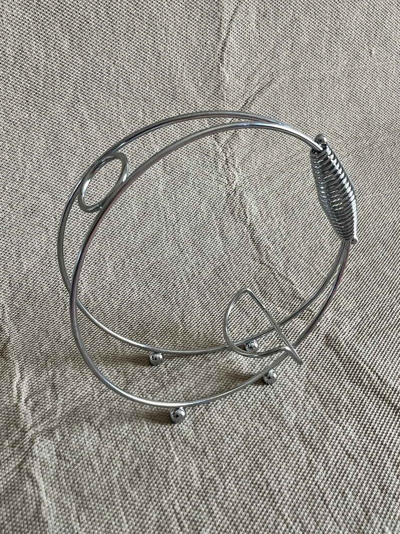 May include: A silver metal wine bottle holder with a circular design. It has a spring-like handle and rests on small, round feet. The design includes decorative oval and curved elements. The holder is made of metal.