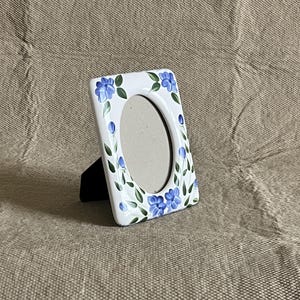 May include: White ceramic photo frame with an oval opening, decorated with blue floral designs and green leaves. The frame is supported by a black stand and is set against a neutral background.