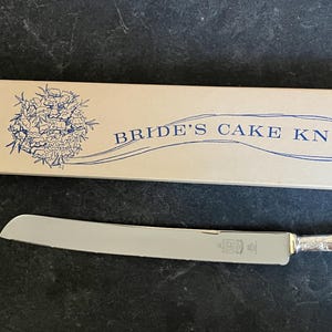 Puede incluir: Un cuchillo plateado para pastel con un mango decorativo descansa junto a su caja. La caja crema presenta ilustraciones florales azules y las palabras "BRIDE'S CAKE KNIFE". El cuchillo tiene un borde dentado y un mango detallado.