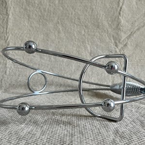 May include: A chrome wine bottle holder with a contemporary design. It has a circular base, curved metal bars, and four small spherical accents. The holder's design is sleek and minimalist, suitable for displaying wine bottles.