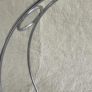 May include: Close-up of a silver metal decorative object with a circular design. The object has a shiny, reflective surface and appears to be made of thin metal rods. The background is a neutral, textured fabric.