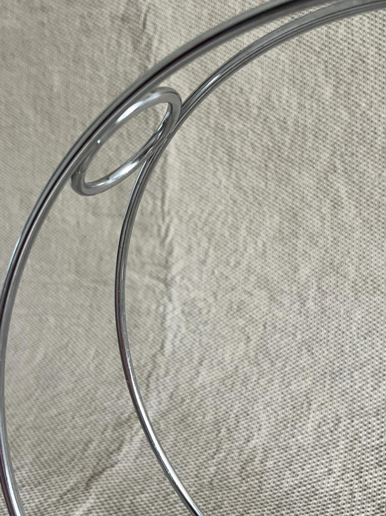 May include: Close-up of a silver metal decorative object with a circular design. The object has a shiny, reflective surface and appears to be made of thin metal rods. The background is a neutral, textured fabric.