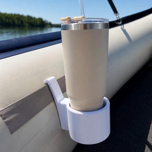May include: A white plastic cup holder attached to the side of an inflatable boat, holding a tall, beige, stainless steel tumbler with a clear lid and a transparent straw. The cup holder features a hook design that secures onto the boat's side.