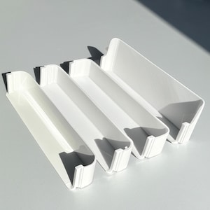May include: Four white rectangular storage containers are arranged in a row. Each container has a slightly different length and a lip on the front edge. The containers are made of a smooth, solid material.