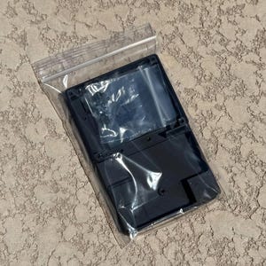 May include: A black plastic electronic device case, sealed in a clear plastic bag. The case has a rectangular shape with a hinged design and a textured surface. The case appears to be empty, with no visible components inside.