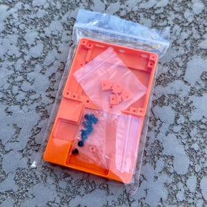 May include: An orange plastic case with a clear plastic bag containing small orange and black hardware pieces. The case has a rectangular shape and is open on one side. The hardware pieces are likely screws and standoffs for assembling a small electronic device.