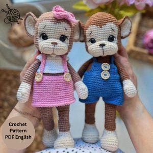May include: Two handmade crochet monkey dolls with brown and cream yarn. The left monkey wears a white top and pink skirt with two buttons. The right monkey wears blue overalls with two buttons. Both have black eyes and embroidered facial features.
