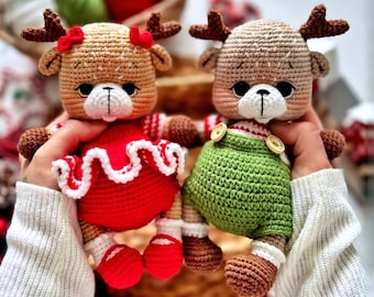 May include: Two handmade crochet reindeer dolls. One doll wears a red dress with white trim and red shoes. The other doll wears green overalls. Both dolls have brown antlers, brown arms and legs, and white faces. The dolls are held by a person wearing a white sweater.