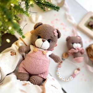 May include: A handmade crochet teddy bear wearing pink overalls with small bear-shaped buttons. The bear has a white muzzle, black eyes, and a small pink bow. A smaller bear and a pacifier are also visible.