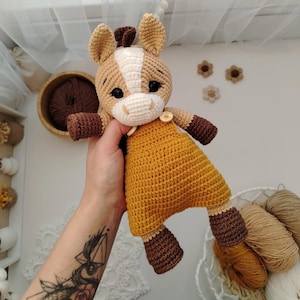 May include: A handmade crochet unicorn doll with a tan and white head, brown mane and hooves, and a mustard yellow dress. The doll has black eyes and a button closure on the dress. The unicorn is a soft toy.
