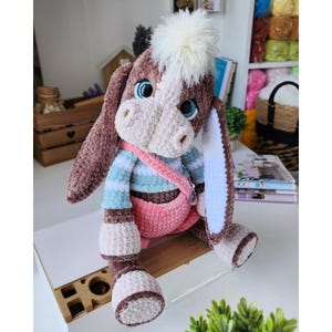 May include: A handcrafted plush donkey toy with a white and brown face, blue eyes, and long ears. It is dressed in a striped blue and white shirt, pink overalls, and a matching pink shoulder bag. The donkey has a fluffy white pom-pom on its head.