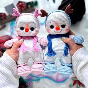 Op de afbeelding: Twee handgemaakte gehaakte sneeuwpoppen. De ene sneeuwpop draagt een roze hoed en sjaal, de andere een blauwe hoed en sjaal. Beide hebben zwarte ogen, een oranje neus en roze wangen. De sneeuwpoppen hebben kleine geweien en dragen bijpassende outfits.