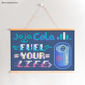 Könnte beinhalten: Kreuzstich-Kunstwerk auf marineblauem Hintergrund mit dem Text "Jojo Cola" und "FUEL YOUR LIFE" in Türkis, Lila und Rosa. Rechts ist eine pixelige Limonadendose. Das Kunstwerk ist in hellem Holz gerahmt und hängt an einer Schnur.