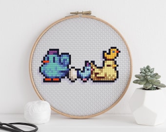 May include: A cross-stitch embroidery hoop featuring pixelated birds and a duck. The birds are blue, and the duck is yellow. The embroidery is set against a light gray fabric, framed by a wooden hoop. The text "@YellowBeeAtelier" is in the top left corner.