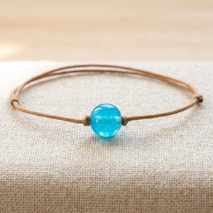 May include: A single round, translucent bright blue bead centred on a thin, light brown cord bracelet. The cord is knotted on both sides of the bead to hold it in place, creating a simple, adjustable band design with a smooth, polished finish.