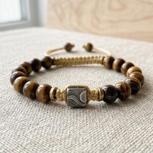 May include: A beaded bracelet featuring polished brown tiger eye stones, a central square metal bead with a swirling gray and black pattern, and tan cord accents. The bracelet includes two small metallic spacers and a sliding knot closure system.