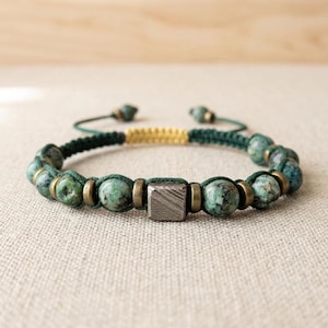 May include: A bracelet featuring round, mottled green and brown stone beads interspersed with small, cylindrical brass-toned metal spacers. A central square, textured dark grey metal bead is positioned at the front, held by dark green cord.