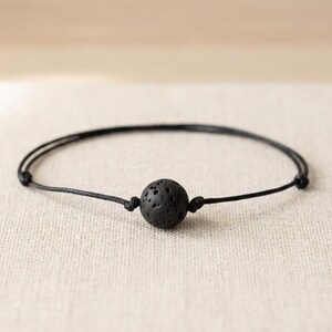 May include: A black bracelet featuring a single spherical lava stone bead with a porous, textured surface. The bead is centered on a thin, black cord that is secured with two small knots on either side of the stone, creating a simple, minimalist design.
