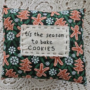 May include: A square decorative pillow with a dark green background featuring gingerbread men, Christmas trees, and snowflakes in shades of brown, white, and green. A stitched fabric patch reads "'tis the season to bake COOKIES."