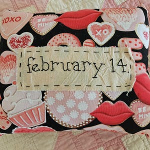 May include: A Valentine's Day themed pillow with a black background, adorned with pink and white hearts, lips, and cupcakes. The phrase "february 14" is embroidered on a fabric patch, adding a personalized touch to this decorative item.