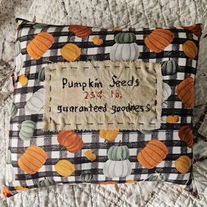 May include: A square decorative cushion featuring a black and white buffalo check pattern with orange, green, and white pumpkin illustrations. A beige fabric patch is stitched to the centre with the text: Pumpkin Seeds, 25¢ lb., guaranteed goodness.