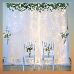 May include: White sheer curtains with twinkling lights create a backdrop for a wedding ceremony. Two white chairs are decorated with floral arrangements.
