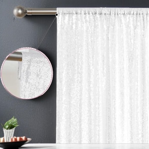 May include: White sheer curtain panel with a textured pattern, hanging from a silver-toned curtain rod. The curtain is partially drawn, revealing the rod and a glimpse of the wall behind. The curtain's fabric appears to be lightweight and allows light to filter through.
