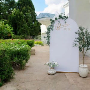 May include: A white arched backdrop with the words "Bride to Be" in gold script, decorated with green vines. A small vase with white flowers and a potted olive tree are in front of the backdrop. The scene is outdoors.