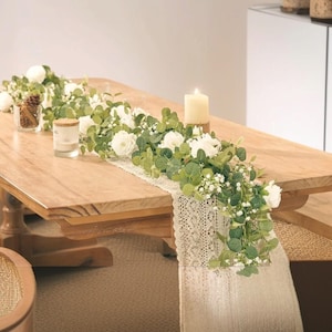 May include: A wooden table is adorned with a floral table runner featuring white roses, green leaves, and delicate white filler flowers. A lit candle and small glass jars with decorative items add to the elegant display.