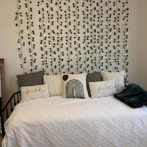 May include: A bed with a white textured comforter and several decorative pillows. The pillows include a gray pillow, a white pillow with a geometric pattern, and a pillow with the word "faith" embroidered on it. Green vines with lights hang on the wall behind the bed.