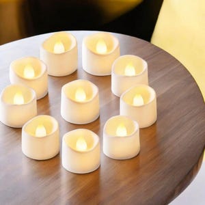 May include: A collection of ten white, battery-operated LED candles with a warm, flickering glow, arranged on a dark wooden surface. The candles are round with a slightly recessed top, mimicking the look of real candles.