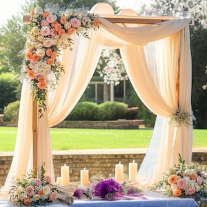 May include: A wooden wedding arch draped with flowing cream-colored fabric and adorned with floral arrangements in shades of peach, pink, and white. The scene includes candles and additional floral bouquets, set against a backdrop of green grass and trees.
