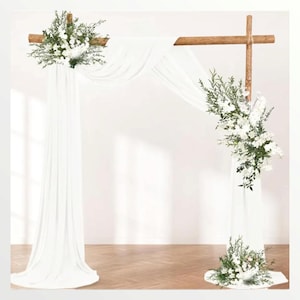 May include: A white fabric backdrop draped over a wooden archway decorated with white flowers and greenery. The archway is made of two wooden beams that are crossed at the top.