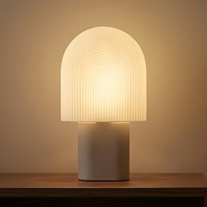 May include: A white table lamp with a cylindrical base and a domed, ribbed shade. The lamp is illuminated, casting a warm glow. It sits on a wooden nightstand, alongside other decorative items.