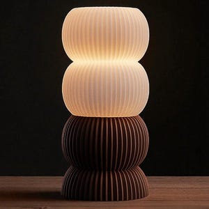 Może przedstawiać: Ułożona lampa stołowa o żebrowanym wzorze. Dwie górne sekcje są białe i podświetlone, a dwie dolne sekcje są brązowe. Lampa stoi na drewnianej powierzchni na ciemnym tle.