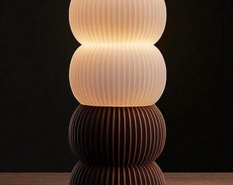 May include: A stacked table lamp with a ribbed design. The top two sections are white and illuminated, while the bottom two sections are brown. The lamp is on a wooden surface against a dark background.