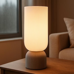 May include: A modern table lamp with a cylindrical, ribbed lampshade and a matching base. The lamp emits a warm, soft light. The base and shade are a neutral color, and the lamp sits on a wooden table.