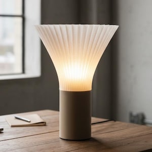 May include: A modern table lamp with a beige cylindrical base and a white, pleated lampshade. The lampshade has a flared, conical shape, and the light glows warmly from within. The lamp sits on a wooden table, with a notebook and pen nearby.