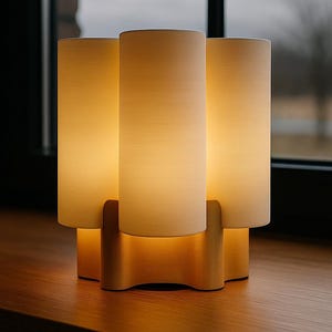May include: A modern table lamp with three cylindrical shades. The shades are a soft white, and the base is made of light-colored wood. The lamp is illuminated, casting a warm glow. The design is minimalist and contemporary.