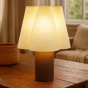 Op de afbeelding: Een moderne tafellamp met een uniek ontwerp. De lamp heeft een cilindrische voet met een kleurverloop van donkerbruin naar lichtbruin, bekroond met een drieledige, crèmekleurige kap. De lamp staat op een houten tafel.