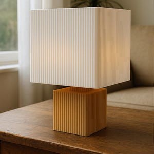May include: A contemporary table lamp featuring a cube-shaped base and lampshade. The base is a warm yellow with vertical grooves, and the lampshade is white with vertical stripes. The lamp is on a dark wooden table.