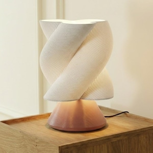 May include: A modern table lamp with a unique twisted shade design. The shade is a light cream color with vertical lines, and the base is a soft pink. The lamp is placed on a wooden nightstand.