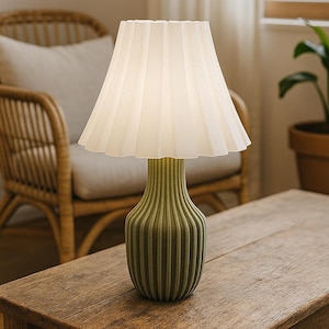 Puede incluir: Una lámpara de mesa decorativa con una pantalla plisada blanca y una base verde acanalada. La lámpara está sobre una mesa de madera, con una silla de mimbre y una planta en maceta en el fondo. Un elemento de decoración del hogar elegante.