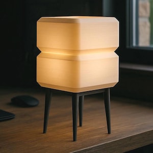 May include: A contemporary table lamp with a stacked, rectangular design. The lamp emits a warm, soft glow and stands on four dark, slender legs. The lamp's dimensions are approximately 25 cm tall and 15 cm wide.