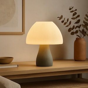 Könnte beinhalten: Eine weiße, pilzförmige Tischlampe mit grünem Fuß steht auf einem Holztisch. Die Lampe ist eingeschaltet und beleuchtet den Raum.